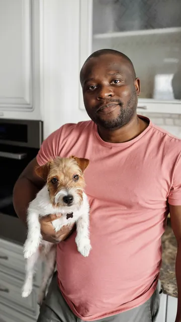 A man holding a small dog in a kitchen, looking at the camera.